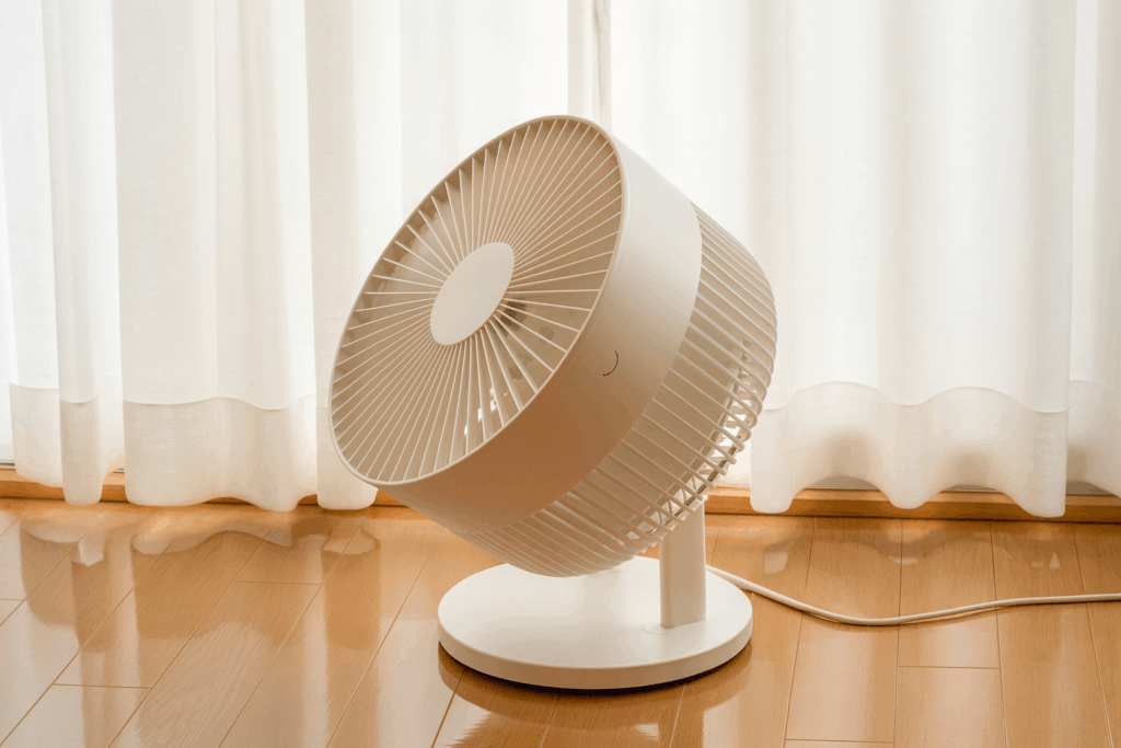 白いサーキュレーターがカーテンの前で稼働している様子。木目の床に光が反射し、ナチュラルで清潔感のある室内空間が広がっている。
