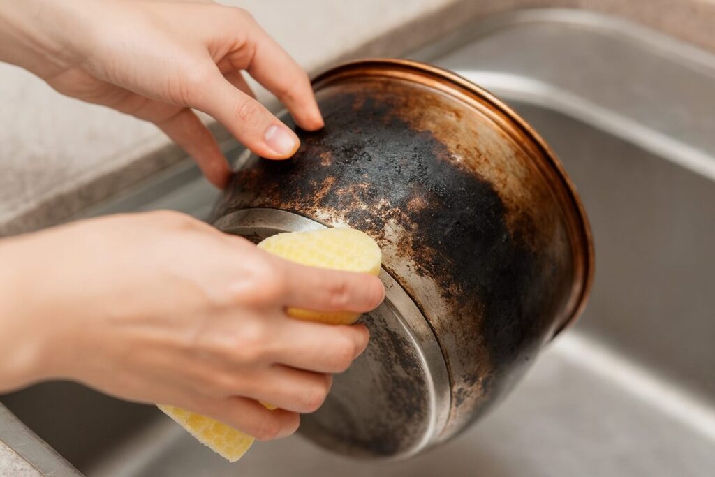 焦げついたステンレス鍋の底面を、スポンジでこすり落としながら洗っている手元を写したイメージ写真。