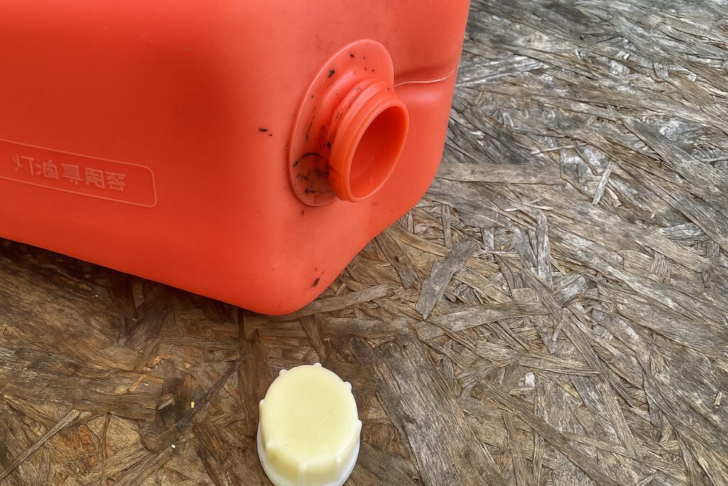 屋外でキャップを外した灯油タンクの注ぎ口を写した写真。赤いタンクの口周りに汚れが付き、白いキャップが地面に置かれている。