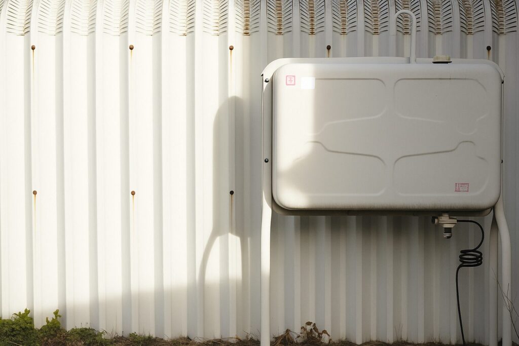 屋外の車庫横に設置された白い灯油タンク。日差しが当たり、タンクと配管の影が壁面に映っている。