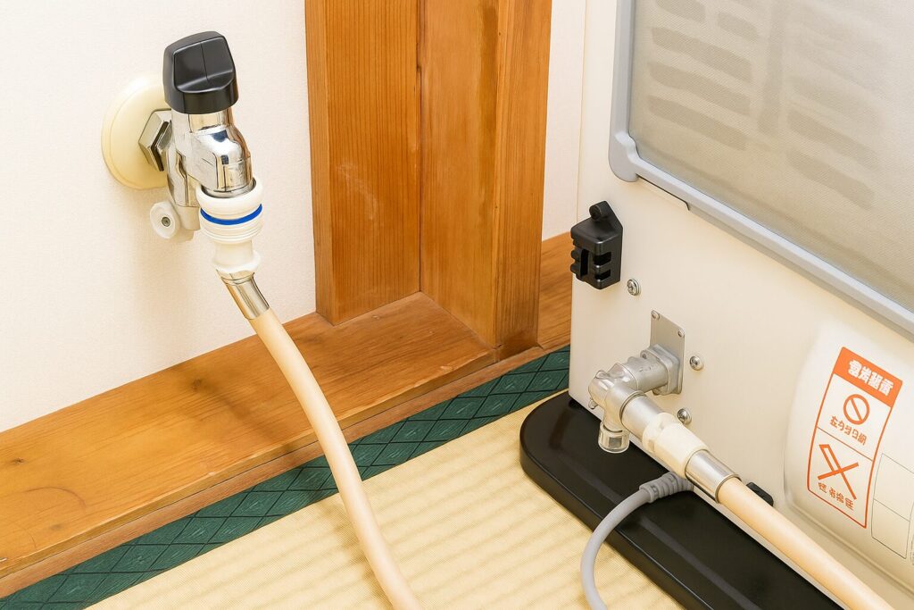 室内の壁に設置されたガス栓と、ガスファンヒーター本体がガスホースで接続されている様子を近距離で写した写真。畳と木枠が見える和室で、接続金具やホースの状態がわかる構図。