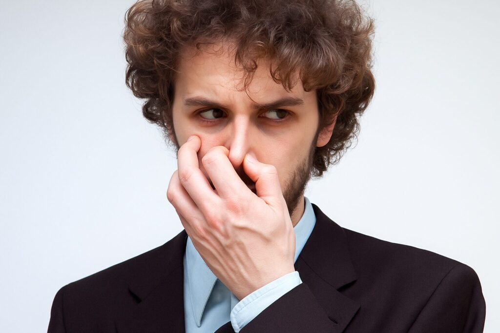 鼻を押さえて臭いを気にしている男性が、不快そうな表情で周囲を見ている様子。背景は白くシンプルで、匂いに反応する瞬間を切り取ったイメージ。