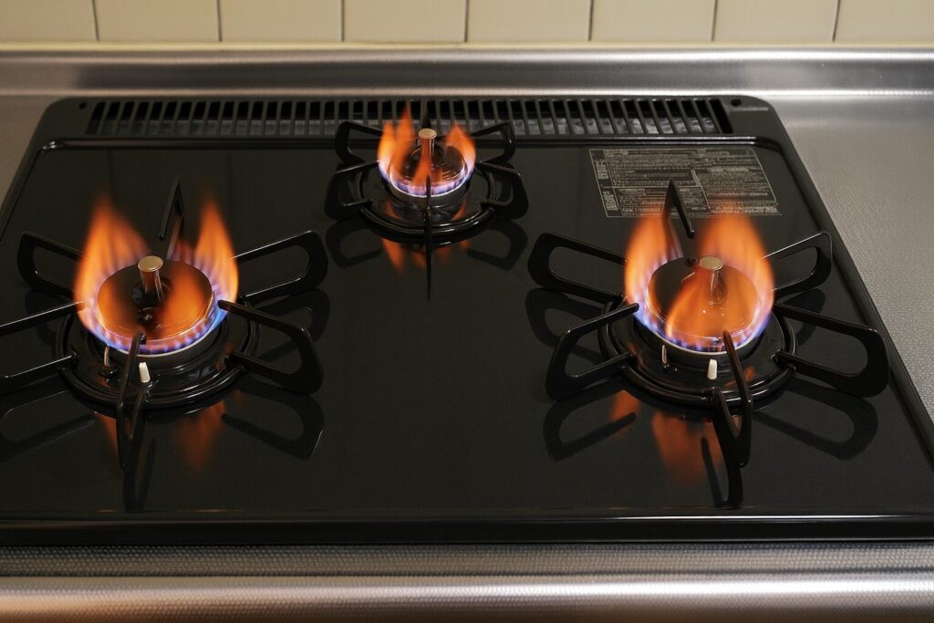ガスコンロの3口バーナーが全て点火し、炎が立ち上がっている様子を正面から写した画像。黒いガラストップが反射し、五徳とバーナーの形状がはっきり見える構図。