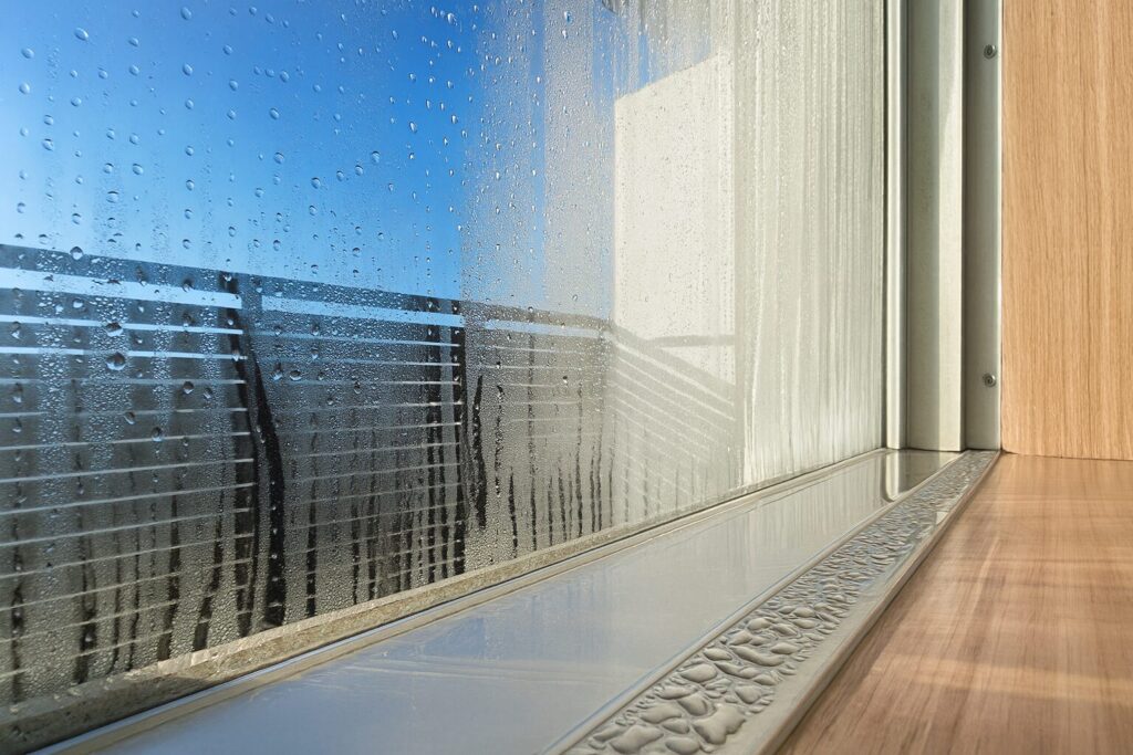 窓ガラスに大量の水滴と結露がつき、窓枠とサッシ部分にも水分が広がっている様子を横長構図で写した画像。屋外の青空と手すりがぼんやりと透けて見え、室内側には光が差し込んでいる。