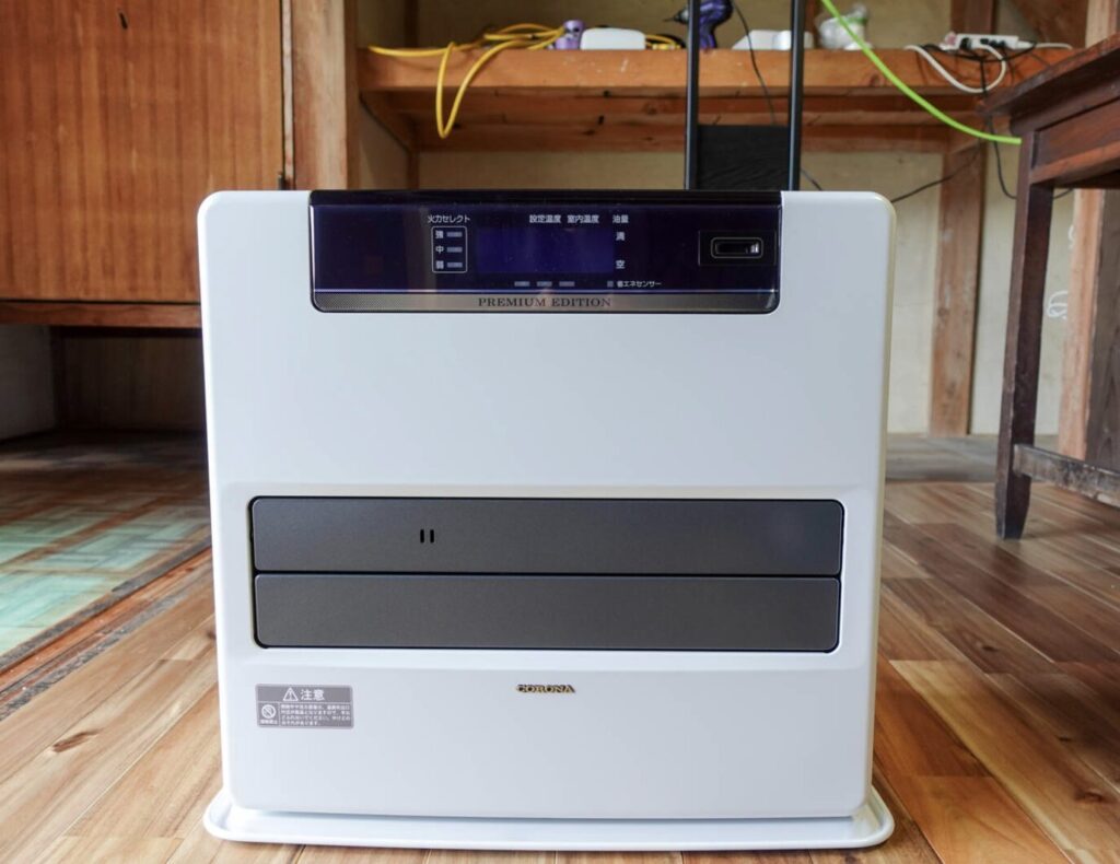 木目調の床の上に設置されたコロナ製の白い石油ファンヒーター。ディスプレイ部分には「PREMIUM EDITION」と表示され、背後には木製の棚とコード類が見える室内の様子。