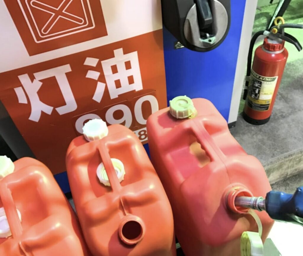ガソリンスタンドで赤いポリタンクに灯油を給油している様子。価格表示の「灯油」看板と消火器が見える夜の給油場面。