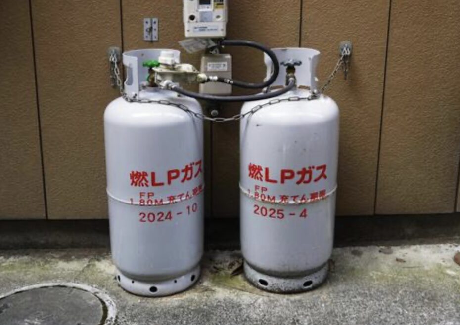 建物の外壁に設置された2本のLPガスボンベ。チェーンで固定され、中央のガスメーターと配管で接続されている様子が写っている。
