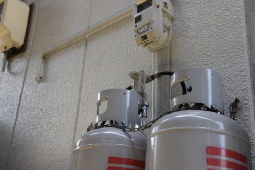 建物の外壁に取り付けられた2本のLPガスボンベ。上部にはガスメーターと配管が接続され、ボンベが鎖で固定されている様子が見える。