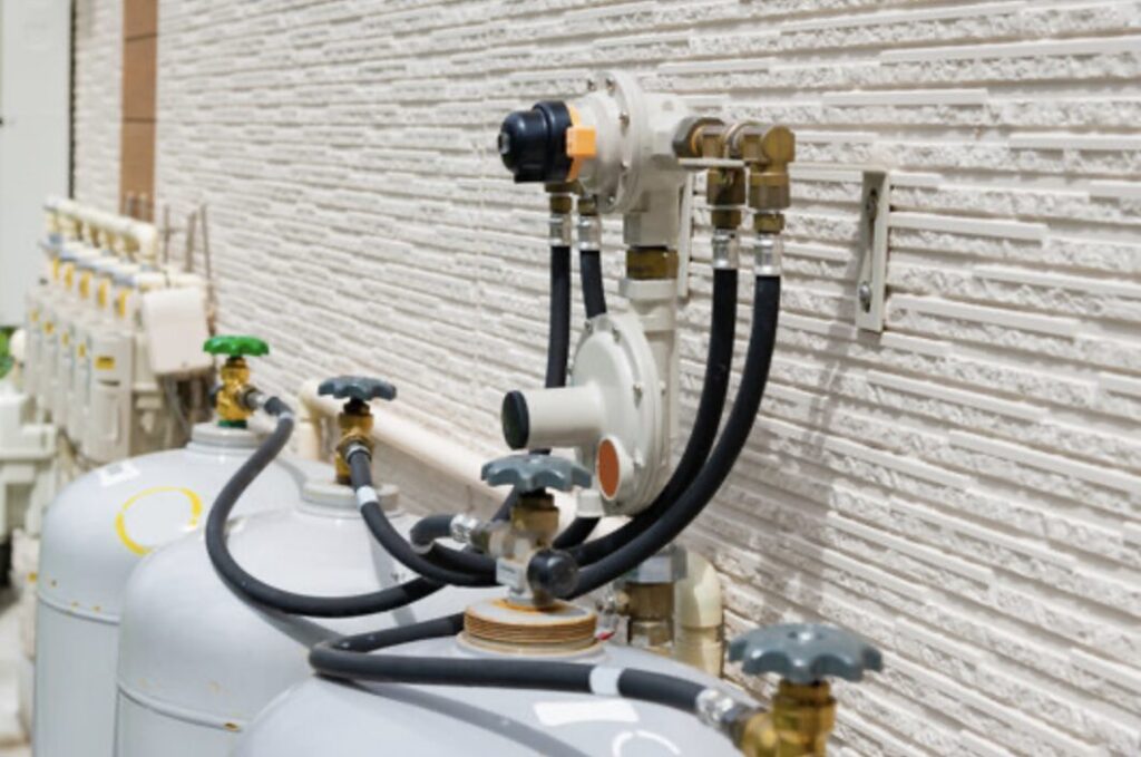 外壁に設置されたLPガスの調整器と配管装置。複数のガスボンベから黒いホースで接続され、圧力を安定させるための調整器が取り付けられている。