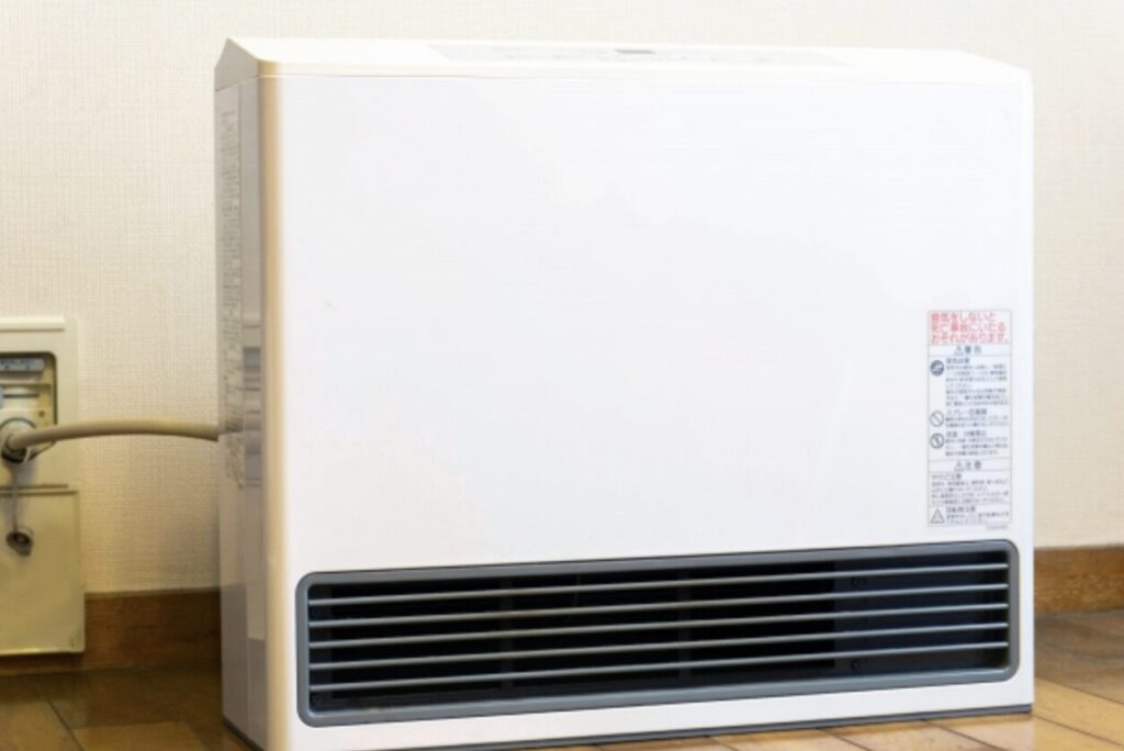 室内に設置された白いガスファンヒーター。ガスコードで壁の元栓につながれ、床付近から温風を送る構造になっている。