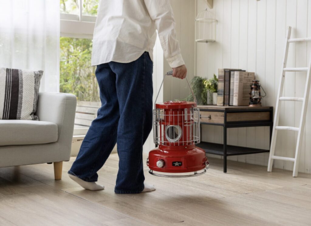 赤いポータブルストーブを持ち運ぶ人の様子。木目調の床と白い壁の室内で、落ち着いた雰囲気のリビング空間が広がっている。