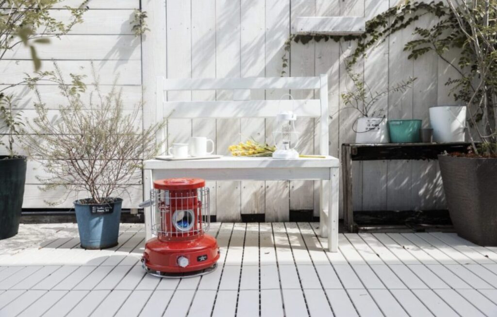 屋外のウッドデッキに置かれた赤いポータブルストーブ。白いベンチや鉢植えが並び、穏やかな日差しの中でくつろぎの空間が演出されている。