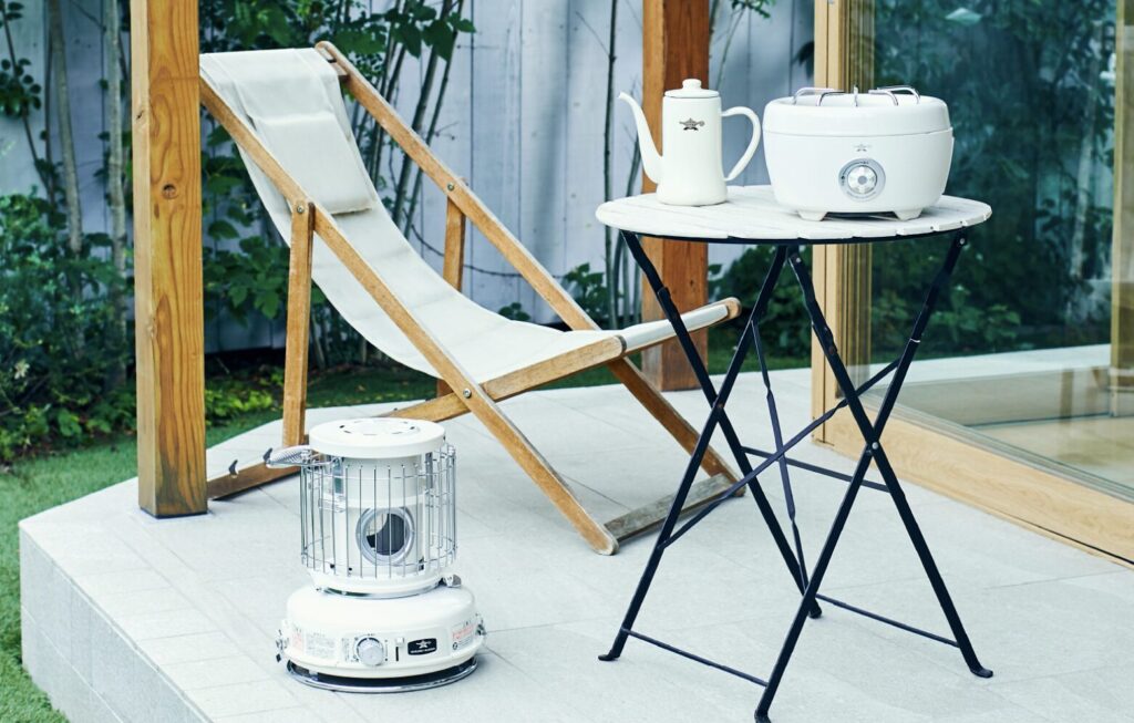 屋外のテラスに置かれた白いポータブルストーブと折りたたみチェア、丸テーブルのある風景。やわらかな自然光が差し込み、リラックスしたカフェのような雰囲気を感じさせる。