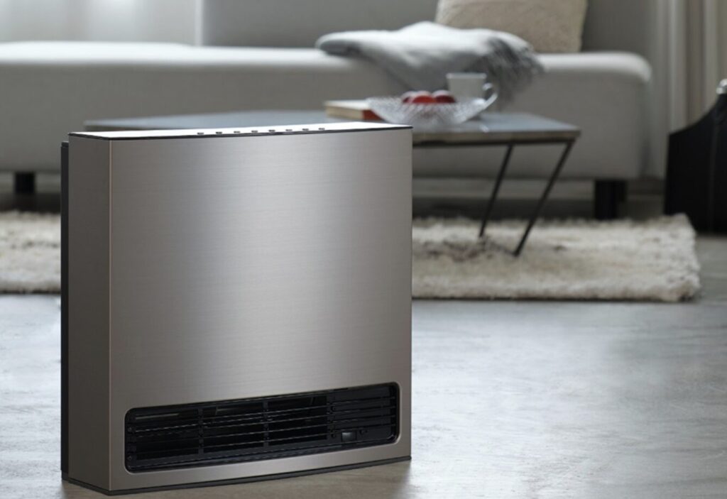 ノーリツ製のガスファンヒーターがリビングに設置されている様子。金属調の本体と下部の送風口が特徴的で、奥にはソファとローテーブルが見える。