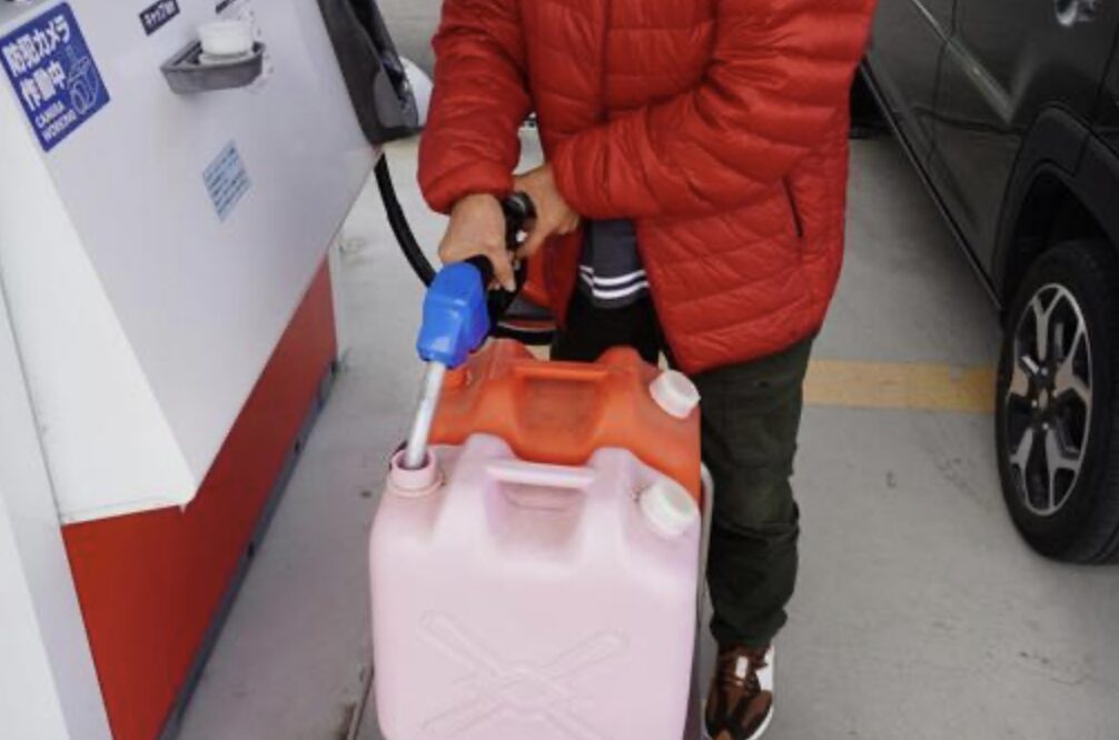 ガソリンスタンドで赤いダウンジャケットの人物が、給油機のノズルを使ってピンク色の灯油ポリタンクに給油している様子