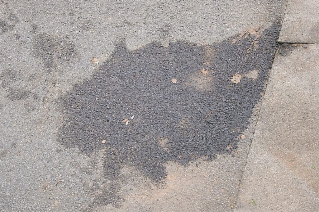 風化したアスファルトの地面に、灯油や油分が染み込んだような濃い色のシミが広がっている様子