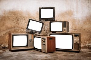 古いブラウン管テレビが複数台、壁際に横一列ではなく段積み状に並べられ、画面が白く光った状態で無機質な室内に置かれている様子