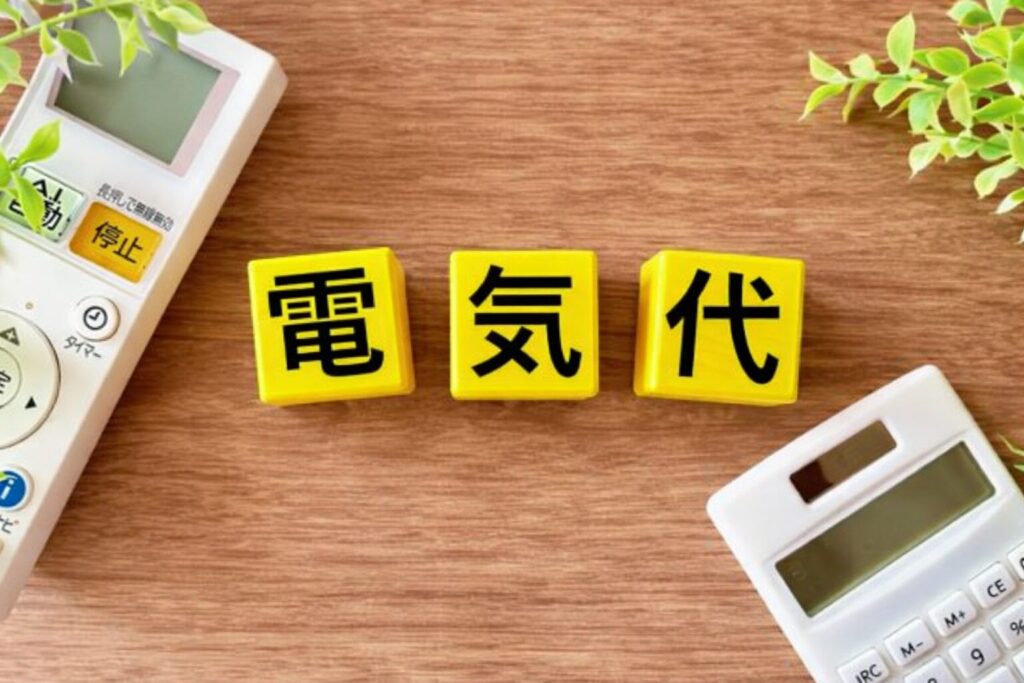 電気代をイメージした黄色のキューブ(電・気・代)が木目のテーブル中央に並び、周囲にエアコンのリモコンと電卓、観葉植物が配置されている様子
