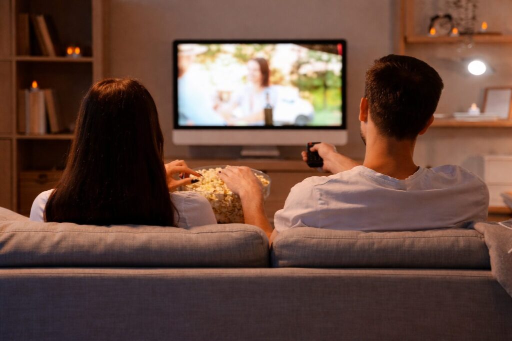 ソファに並んで座る男女が、リモコンを操作しながらテレビ番組を視聴し、中央のボウルに入ったポップコーンをつまんでくつろいでいるリビングの様子