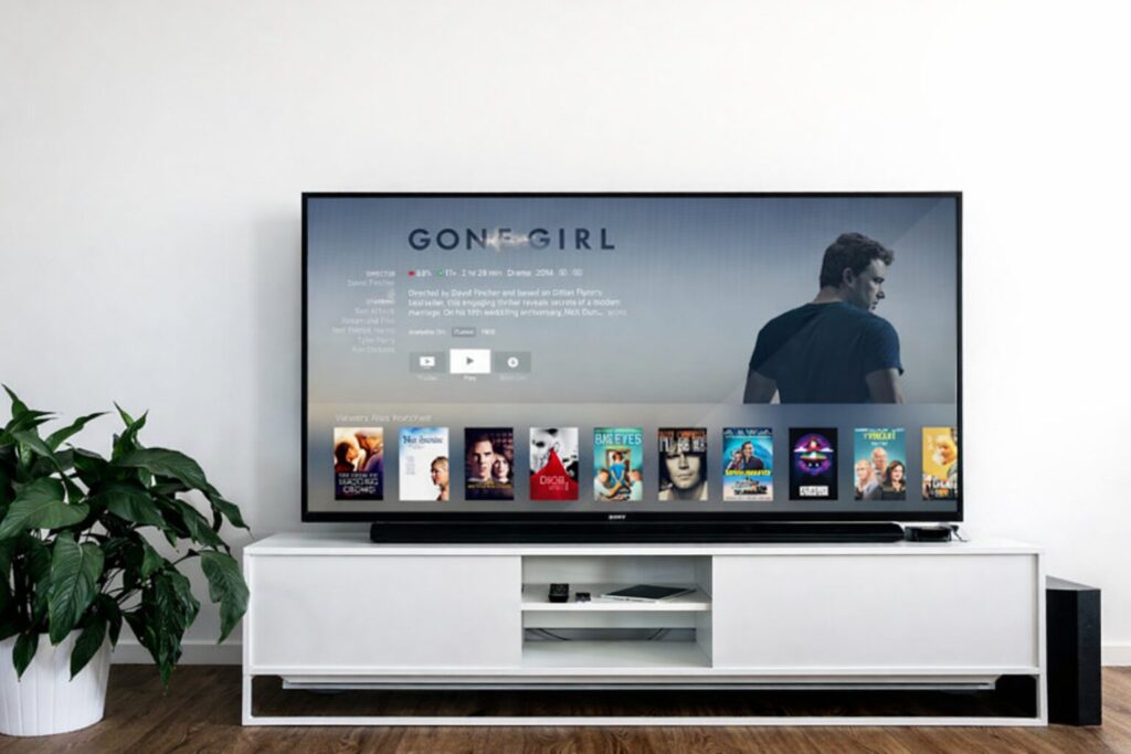 白い壁の前に設置されたテレビ台の上に大型テレビが置かれ、画面には動画配信サービスの作品一覧と映画の再生画面が表示されているリビングの様子。左側には観葉植物、床は木目調でシンプルかつ整った室内空間が広がっている構図。