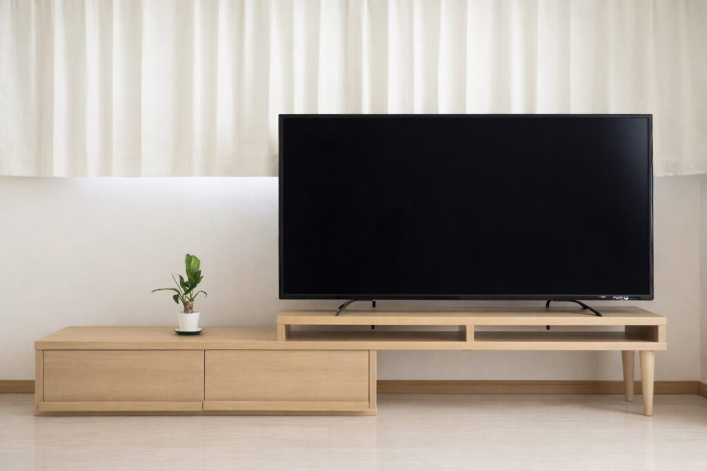 白いカーテンの前に、明るい木目のテレビボードと薄型テレビが設置され、左側に小さな観葉植物が置かれたシンプルで整理されたリビング空間の様子