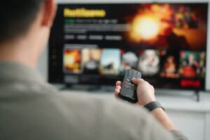 テレビ画面に向かって座り、手にしたリモコンで番組を操作しようとしている人物の後ろ姿と、画面上に並ぶ映像コンテンツがぼかして映る様子