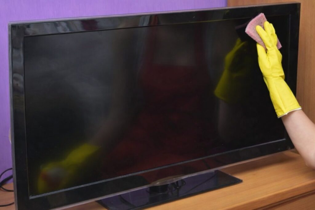 黄色いゴム手袋を着けた手が、布でテレビ画面の汚れを拭き取っている様子。