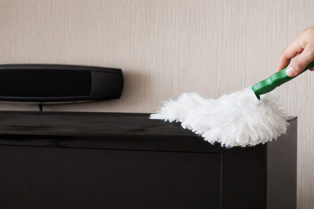 白いモップでテレビ台の上部をやさしく掃除している手元の様子。黒いテレビ台とスピーカーが並び、ホコリを取り除いている場面が横から写っている。