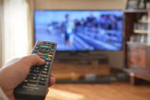 テレビに向けてリモコンを操作している手元の様子。背景にはスポーツ番組が映るテレビ画面がぼかして表示されている。