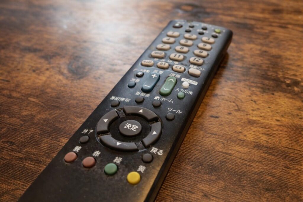 木目のテーブルの上に置かれた日本語表記のテレビ用リモコンを斜め上から捉えた写真。数字ボタンや決定ボタンが見え、使用感のある質感が伝わる構図。