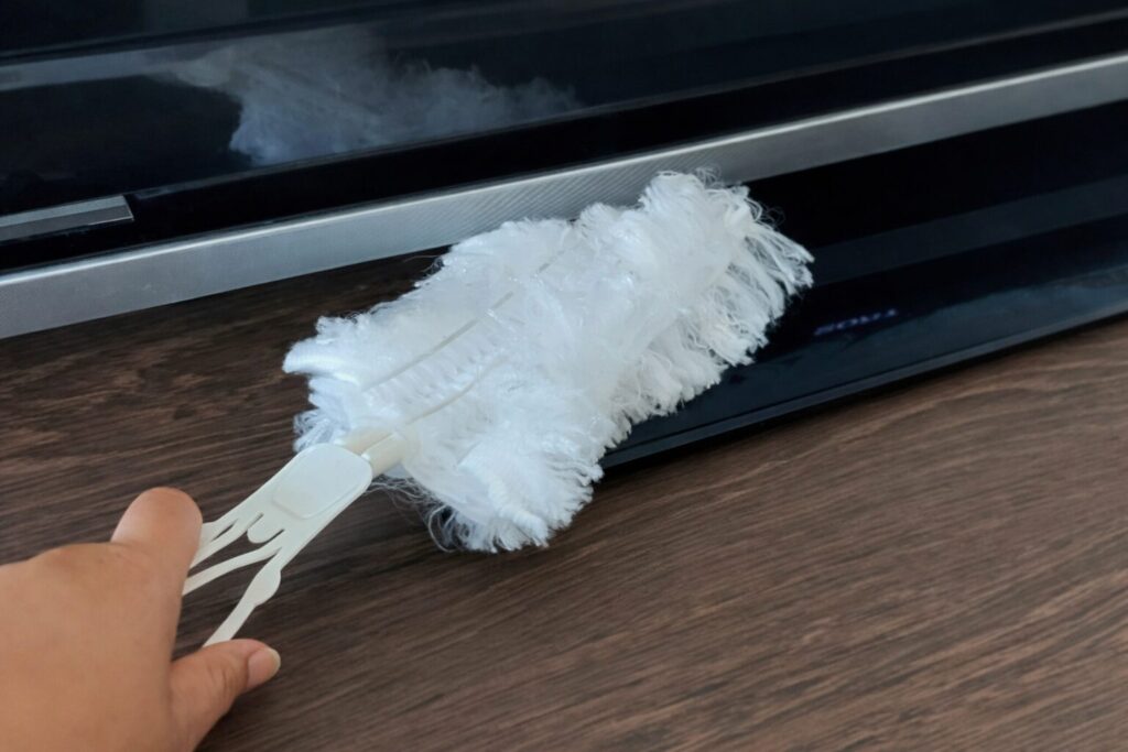 テレビ台の上に置かれた薄型テレビの下部フレーム部分を、白いハンディモップでやさしくホコリ取りしている手元の様子