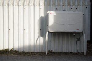 屋外の白い外壁に設置された据え置き型の灯油タンクを正面から捉えた様子。金属製の脚で地面から浮かせて固定され、下部には配管とホースが接続されている。