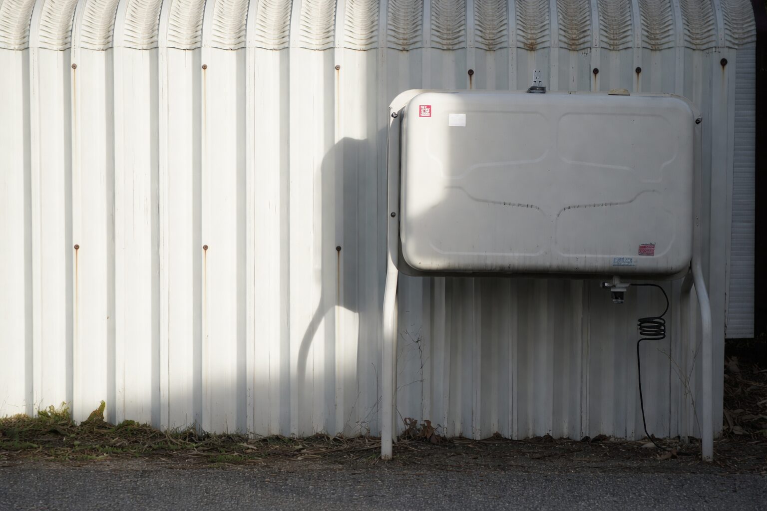 屋外の白い外壁に設置された据え置き型の灯油タンクを正面から捉えた様子。金属製の脚で地面から浮かせて固定され、下部には配管とホースが接続されている。