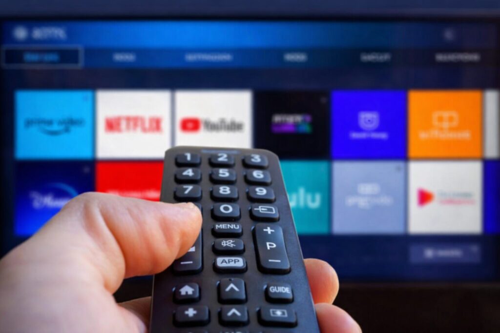 テレビ画面に向けてリモコンを操作する手元の様子。背景には動画配信サービスのアイコンが並び、リモコンのボタンに指をかけて操作している状況がわかる。