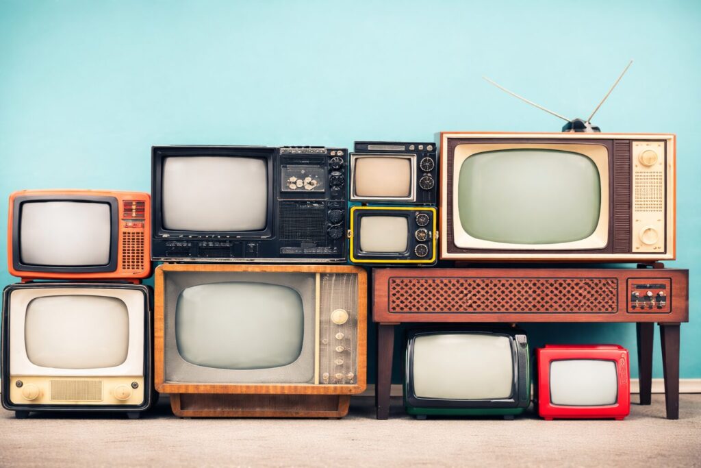 複数台のレトロなブラウン管テレビが、ターコイズブルーの壁を背景に横一列へ重なるように配置されている様子。木目調やカラフルな筐体、ダイヤル式操作部やアンテナ付きの機種など、年代ごとのデザインの違いがわかる構図。