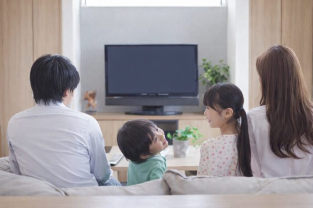 テレビの前に置かれたソファに家族4人が並んで座り、画面のついていないテレビを囲んでくつろいでいるリビングの様子。中央の子どもが振り返って家族の顔を見上げ、自然光が差し込む落ち着いた室内空間が伝わる構図。