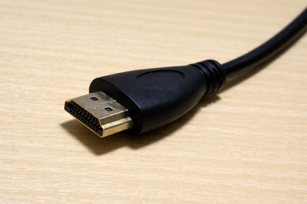 HDMIケーブルの先端コネクタ部分を接写した様子。黒い樹脂製のコネクタと金属端子が、明るい木目の机の上に横向きで置かれている。