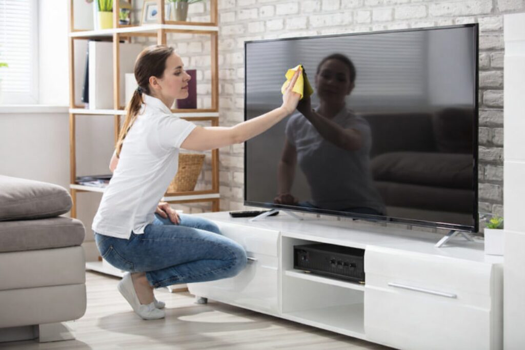テレビ画面を傷つけないように、黄色のクロスで優しく拭き掃除をしている女性が、明るいリビングで大型テレビの前にしゃがんでいる様子