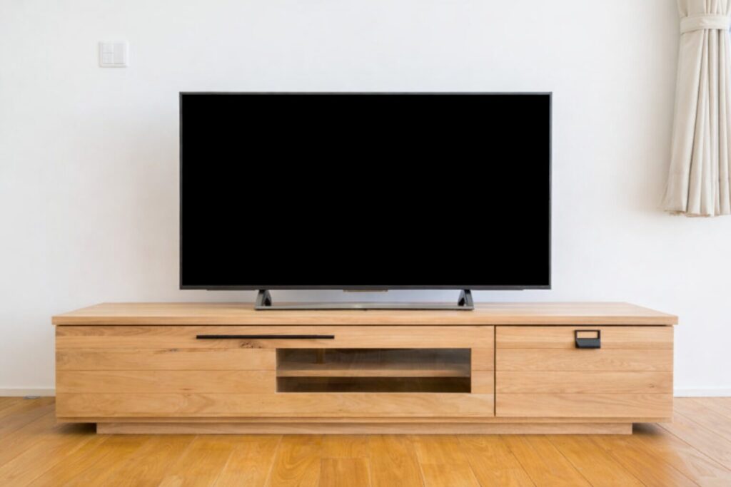 白い壁の前に設置された木製テレビボードの上に、電源オフ状態の薄型テレビが中央に置かれているシンプルなリビングの様子