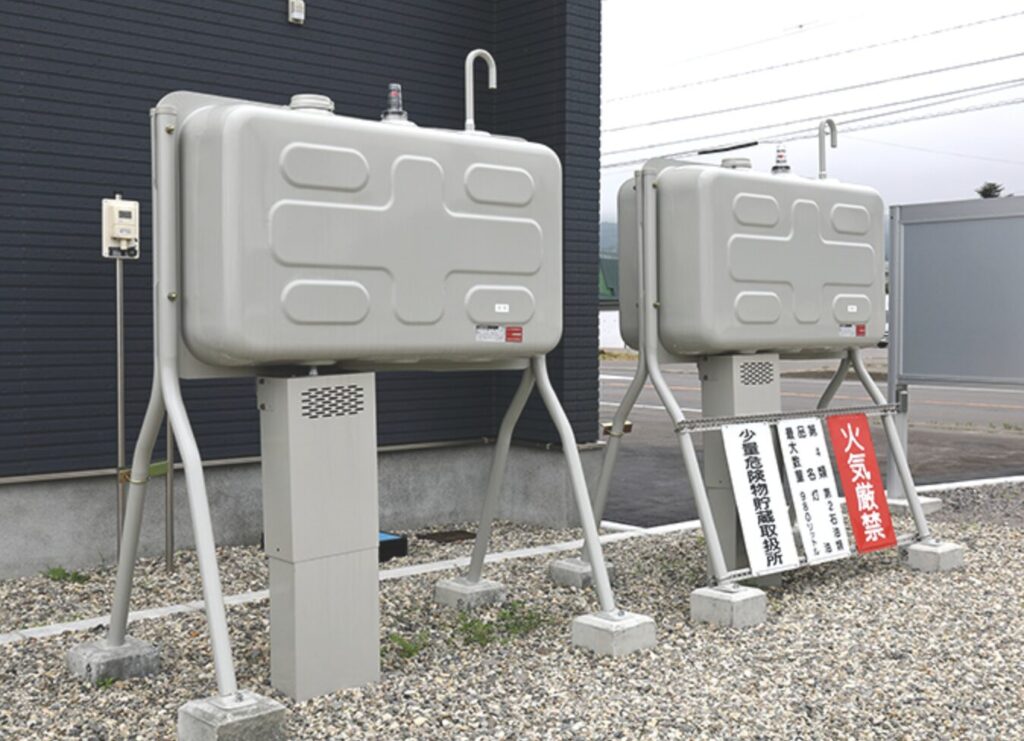 屋外に設置された二基の灯油ホームタンクが金属製の架台に載って並んでいる様子。足元にはコンクリートブロックがあり、手前には火気厳禁の注意看板が立て掛けられている。