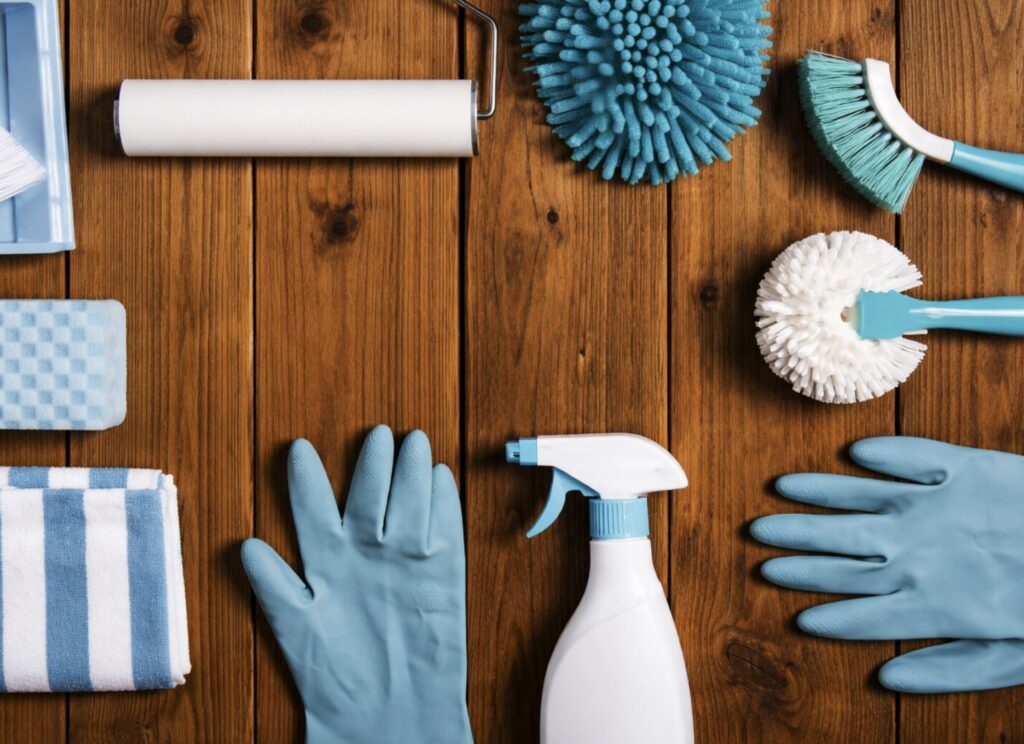 清掃用品を上から見下ろした構図で、木目のテーブルの上に青と白を基調としたブラシ、スポンジ、ゴム手袋、スプレーボトル、ローラーなどが整然と並んでいる様子を表した横長のイメージ。