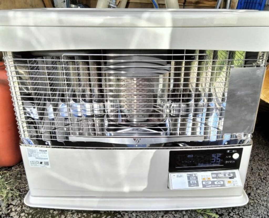 屋外に置いてあるサンポット製の石油ストーブ本体を正面から撮影した様子。前面のガード越しに燃焼部と内部構造が見え、右下には操作パネルが配置されている。