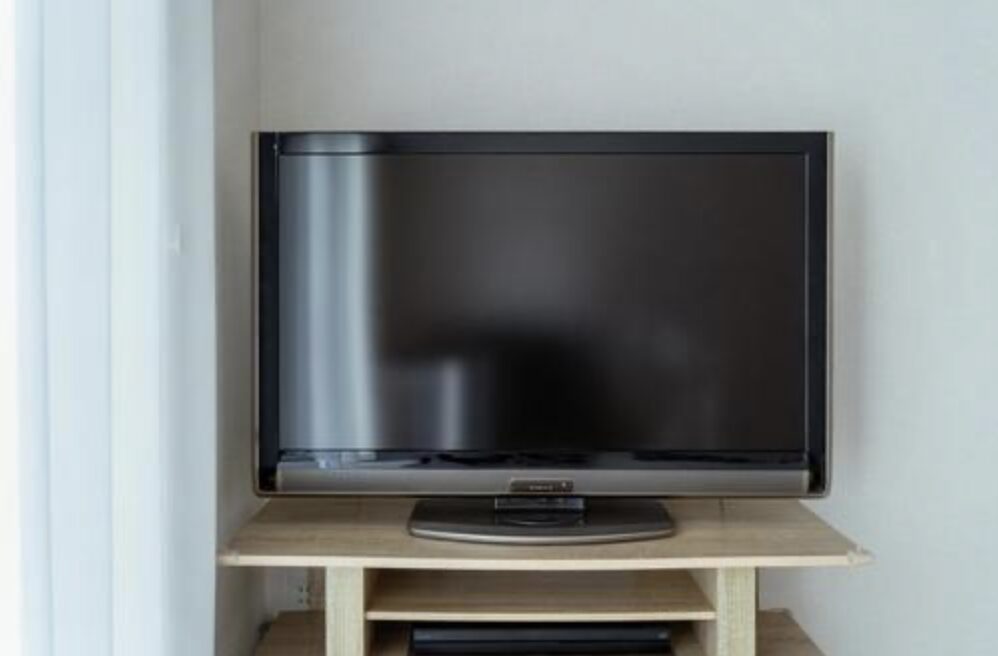 白い壁に囲まれたスペースに設置されたテレビ台の上に、電源が入っていない状態の薄型テレビが正面から置かれている様子