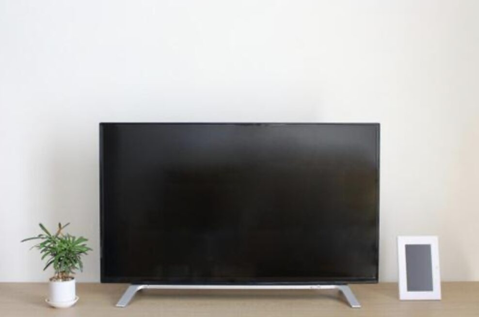 木目のテーブルの上に置かれた薄型テレビを正面から撮影した構図で、左側に小さな観葉植物、右側にフォトフレームが並び、電源が入っていない黒い画面が目立つ室内の様子