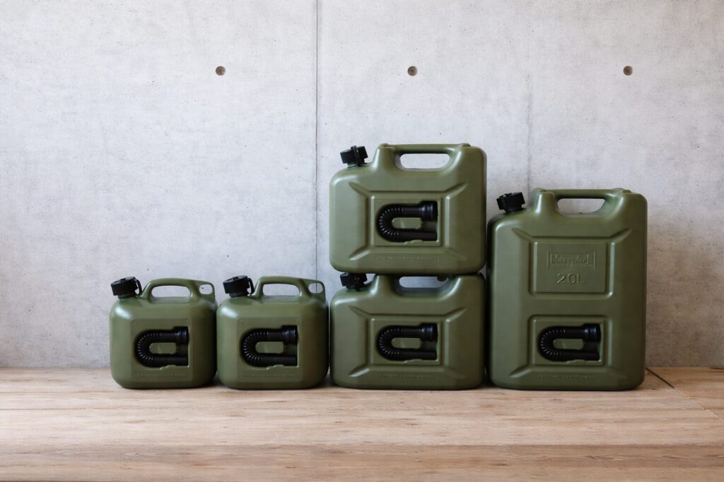 オリーブグリーン色の灯油用ポリタンクが5個並べられている様子。容量の異なるポリタンクがコンクリート壁の前に配置され、床は木製で、各タンクには黒いキャップと注ぎ口が付いている。
