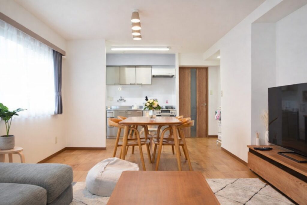 白を基調とした明るいリビングダイニングで、木製のダイニングテーブルと椅子が中央に配置され、奥にはシンプルなキッチン、右側にはテレビボードとテレビが見えるすっきりとした室内の様子。