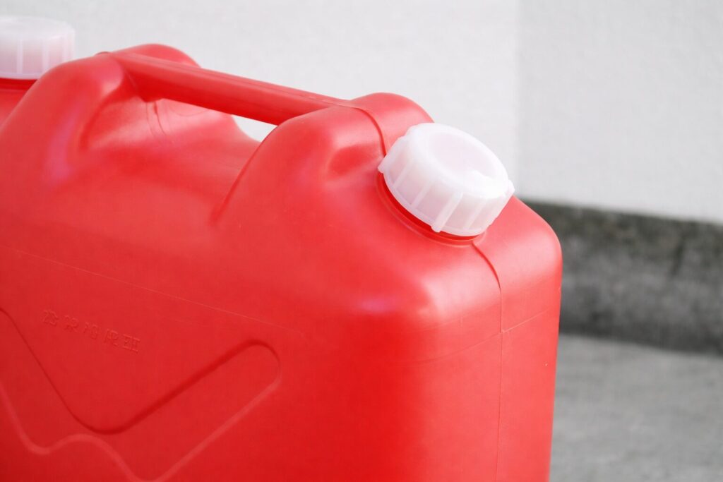 赤色のポリタンクを横から捉えた構図。持ち手一体型の本体上部に、白色のスクリューキャップがしっかりと装着されており、屋外のコンクリート付近に置かれている様子がわかる。