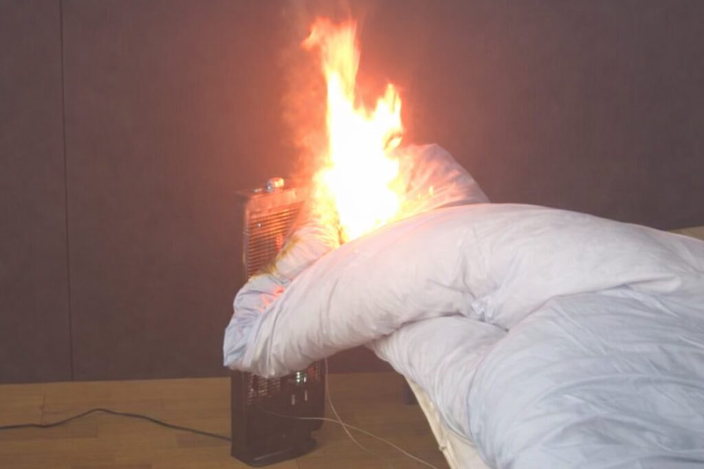 電気ストーブの前面に掛かった布団が熱で発火し、炎が大きく立ち上がっている危険な状況を示す様子。