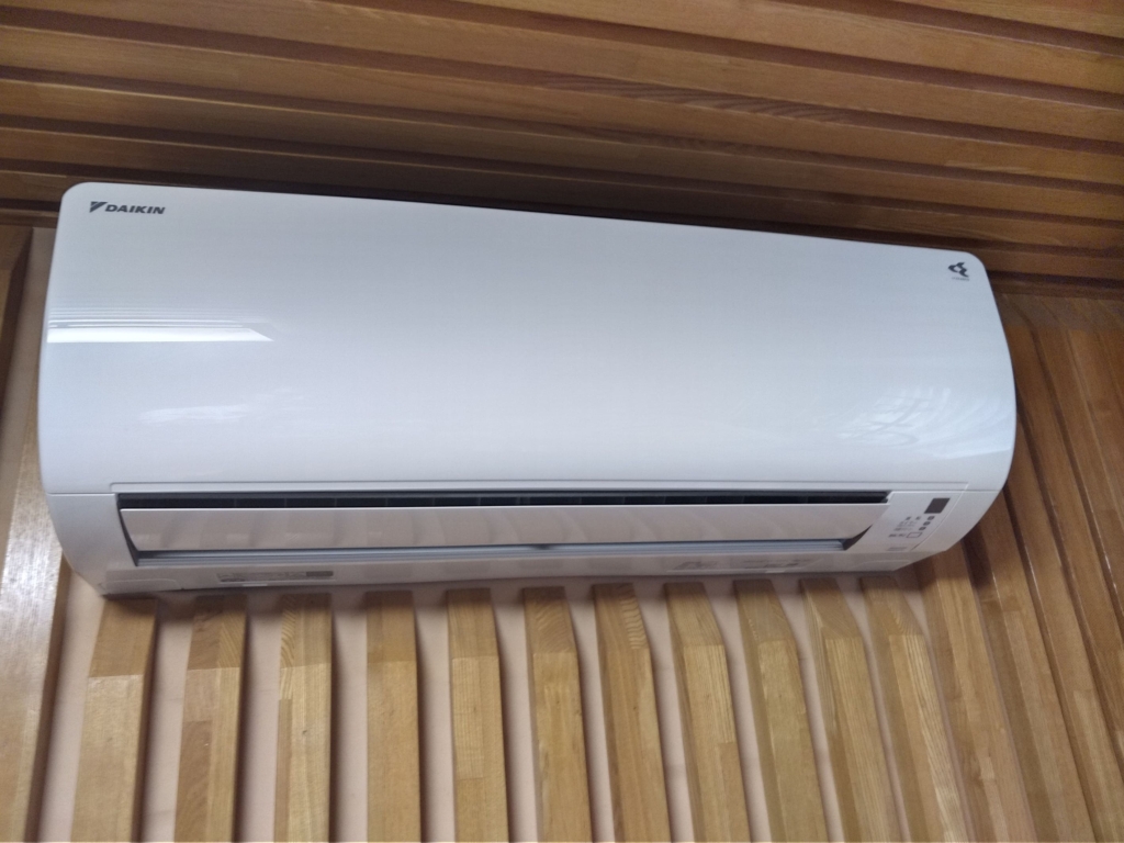 天井近くの木目調の壁に設置されたダイキン製の壁掛けエアコン室内機。運転停止中で吹き出し口が閉じた状態。