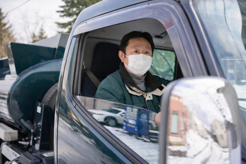 灯油配送用タンクローリーの運転席で、作業着とマスクを着用した運転手が窓越しに前方を確認している様子。車両後方には円筒形の灯油タンクが搭載され、サイドミラーには雪の残る住宅街の景色が映り込んでいる構図。